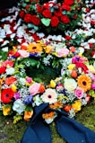 A colorful floral arrangement with a variety of flowers including roses, gerbera daisies, and tulips. Red rose petals are scattered in the background, enhancing the vibrant display on a bed of moss. A dark ribbon is incorporated into the arrangement, suggesting a formal or somber occasion.