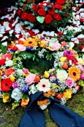 A colorful floral arrangement with a variety of flowers including roses, gerbera daisies, and tulips. Red rose petals are scattered in the background, enhancing the vibrant display on a bed of moss. A dark ribbon is incorporated into the arrangement, suggesting a formal or somber occasion.