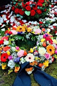 A colorful floral arrangement with a variety of flowers including roses, gerbera daisies, and tulips. Red rose petals are scattered in the background, enhancing the vibrant display on a bed of moss. A dark ribbon is incorporated into the arrangement, suggesting a formal or somber occasion.