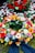 A colorful floral arrangement with a variety of flowers including roses, gerbera daisies, and tulips. Red rose petals are scattered in the background, enhancing the vibrant display on a bed of moss. A dark ribbon is incorporated into the arrangement, suggesting a formal or somber occasion.