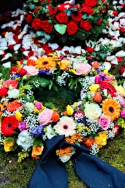 A colorful floral arrangement with a variety of flowers including roses, gerbera daisies, and tulips. Red rose petals are scattered in the background, enhancing the vibrant display on a bed of moss. A dark ribbon is incorporated into the arrangement, suggesting a formal or somber occasion.