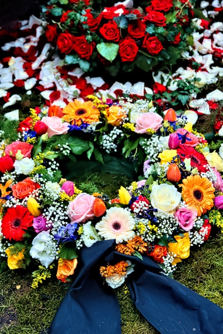 A colorful floral arrangement with a variety of flowers including roses, gerbera daisies, and tulips. Red rose petals are scattered in the background, enhancing the vibrant display on a bed of moss. A dark ribbon is incorporated into the arrangement, suggesting a formal or somber occasion.