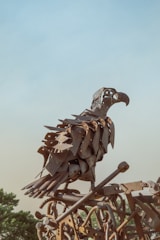 A sculpture of a bird made from various metal parts, giving it a rustic and industrial look. The artwork is perched against a backdrop of a clear blue sky, with some greenery visible in the lower part of the image.