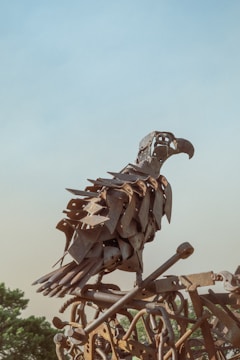 A sculpture of a bird made from various metal parts, giving it a rustic and industrial look. The artwork is perched against a backdrop of a clear blue sky, with some greenery visible in the lower part of the image.