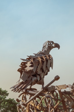 A sculpture of a bird made from various metal parts, giving it a rustic and industrial look. The artwork is perched against a backdrop of a clear blue sky, with some greenery visible in the lower part of the image.