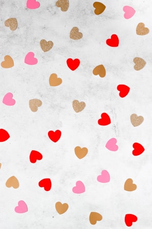 A pattern of various heart shapes scattered across a light gray textured background. The hearts are in shades of red, pink, gold glitter, and brown, creating a playful and decorative arrangement.