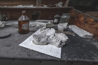 A dusty workspace features dental molds prominently placed on a piece of paper. Various bottles, some with labels, are scattered across the surface. The background contains a wooden compartment, adding to the aged, neglected atmosphere.