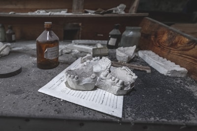 A dusty workspace features dental molds prominently placed on a piece of paper. Various bottles, some with labels, are scattered across the surface. The background contains a wooden compartment, adding to the aged, neglected atmosphere.