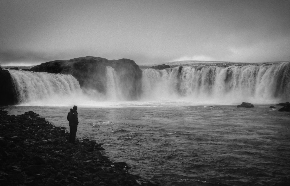 grayscale photo of man standing near waterfalls