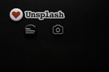 A black textured background with four small objects arranged on it. There is a white badge with a red heart symbol, a cutout spelling 'Unsplash', a round black button with white text saying 'Make something awesome.', and a small camera icon.