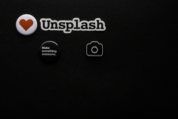 A black textured background with four small objects arranged on it. There is a white badge with a red heart symbol, a cutout spelling 'Unsplash', a round black button with white text saying 'Make something awesome.', and a small camera icon.
