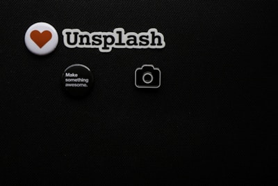 A black textured background with four small objects arranged on it. There is a white badge with a red heart symbol, a cutout spelling 'Unsplash', a round black button with white text saying 'Make something awesome.', and a small camera icon.