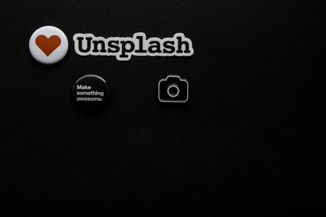 A black textured background with four small objects arranged on it. There is a white badge with a red heart symbol, a cutout spelling 'Unsplash', a round black button with white text saying 'Make something awesome.', and a small camera icon.
