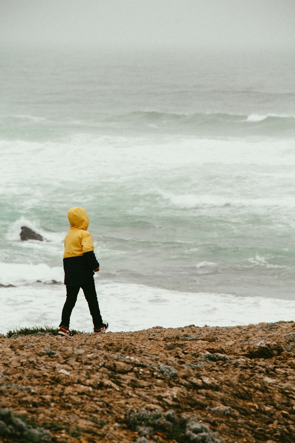 Mann in gelbem Kapuzenpulli, der tagsüber auf einem braunen Felsen in der Nähe des Meeres steht