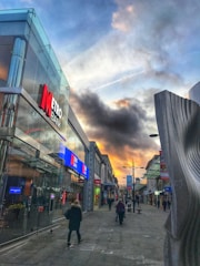 A city street scene with a bank on the left, reflecting glass windows, and several pedestrians walking along the sidewalk. The sky features dramatic clouds with hints of sunlight, and there is a modern, abstract sculpture on the right side.
