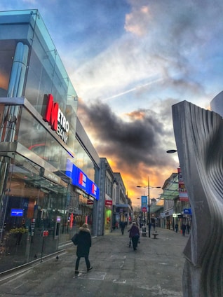 A city street scene with a bank on the left, reflecting glass windows, and several pedestrians walking along the sidewalk. The sky features dramatic clouds with hints of sunlight, and there is a modern, abstract sculpture on the right side.
