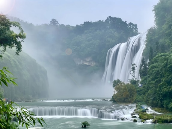 A majestic waterfall cascades down a lush green mountainside into a misty pool below, surrounded by dense forest and vegetation. The scene is enveloped in gentle mist and soft sunlight, creating a serene atmosphere.