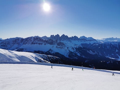 Four skiers descend a snowy slope under a clear blue sky. The sun shines brightly above a jagged range of mountain peaks in the background.