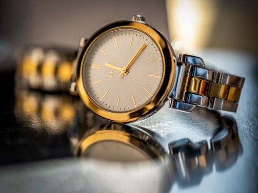 A close-up view of a luxurious wristwatch resting on a reflective surface. The watch features a metallic silver and gold-tone design, with a sleek round face and minimalist hour markers.