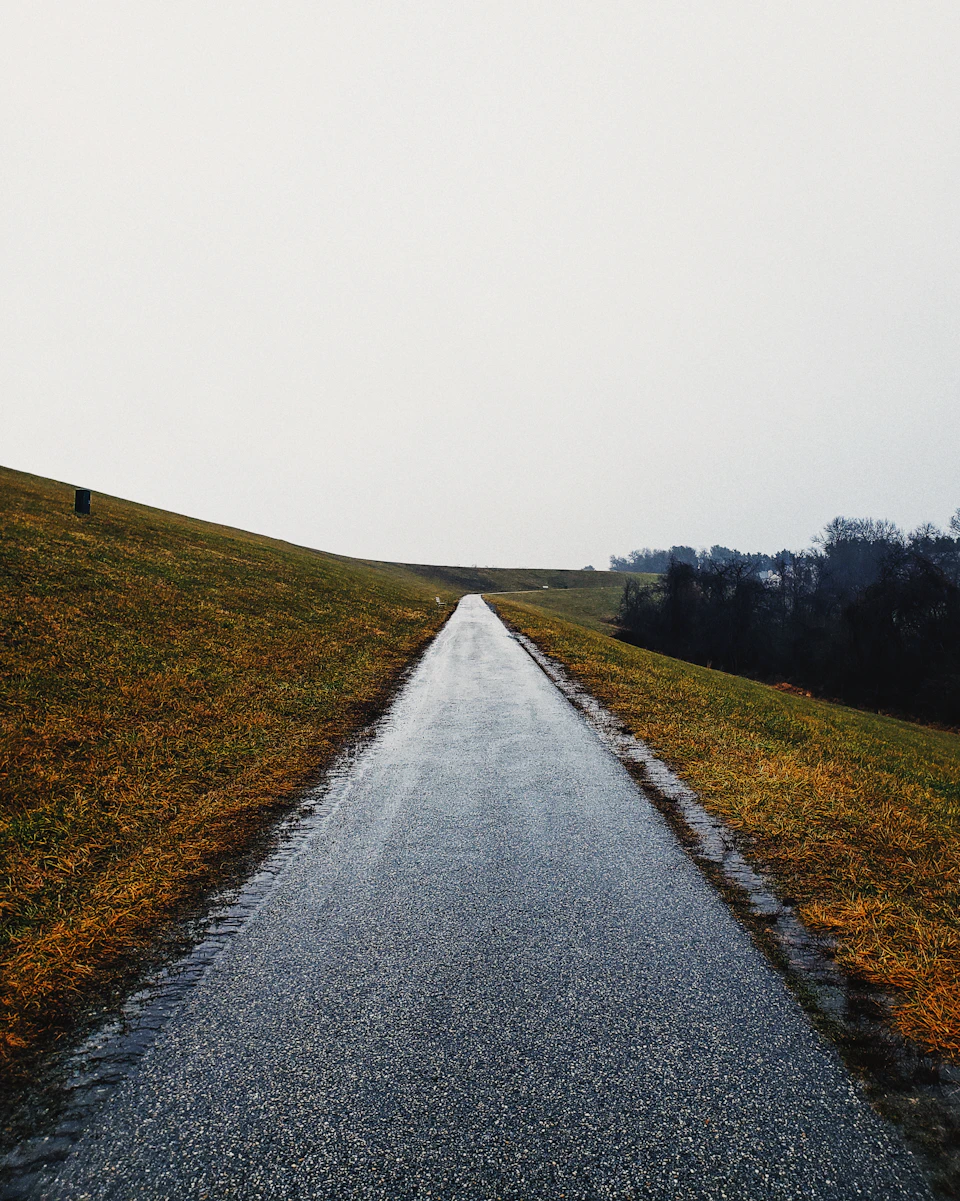 Long, straight road cuts through a grassy field on a cloudy, rainy day outdoors