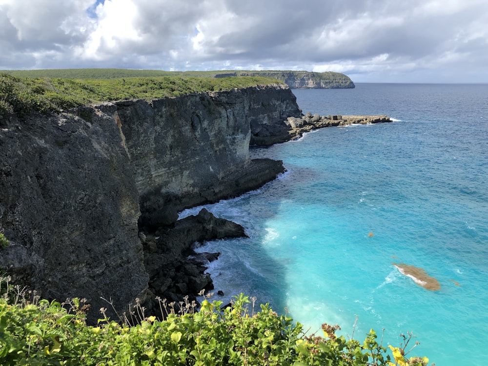 montagna rocciosa verde e marrone accanto al mare blu sotto il cielo nuvoloso blu e bianco durante il giorno