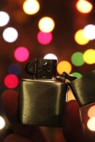 A metal lighter is being held up against a backdrop of colorful, out-of-focus lights creating a festive bokeh effect. The lighter is open, revealing the ignition mechanism, and is prominently featured in the foreground.