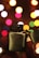 A metal lighter is being held up against a backdrop of colorful, out-of-focus lights creating a festive bokeh effect. The lighter is open, revealing the ignition mechanism, and is prominently featured in the foreground.
