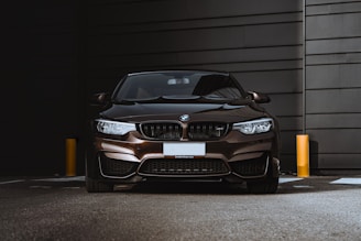A sleek, dark-colored BMW car is parked against a modern, industrial-looking backdrop. The lighting highlights the glossy surface of the vehicle, emphasizing its design features.
