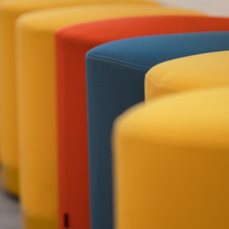 A close-up of a row of colorful, curved seating stools in a pattern. The colors include yellow, red, and dark teal, providing a modern and vibrant feel.