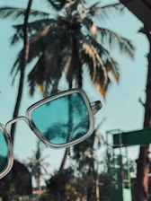 A pair of sunglasses with blue-tinted lenses is held up against a background of tall palm trees, creating a tropical atmosphere. The palm trees appear slightly blurred, contrasting with the sharp focus of the sunglasses.