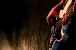 A hand is playing a bass guitar with a focus on the strings and body of the instrument. The background is dark and out of focus, highlighting the musician's hand and the guitar.