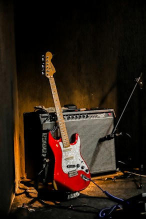 A red electric guitar leans against a black amplifier in a dimly lit room. The guitar features a white pickguard and multiple control knobs. A microphone stand is visible to the side, suggesting a musical setup. The scene is set against a textured dark wall, with some shadow play in the lighting.