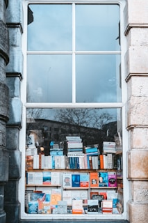 A large window display features an array of books neatly stacked and arranged on shelves. The window reflects a cloudy sky and leafless trees outside. The books are of various sizes and colors, with some titles visible toward the bottom.