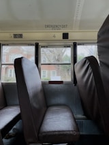 The image features the interior of a school bus, focusing on two rows of brown seats positioned against a window. Outside, there is a view of red brick houses and leafless trees. Above the seats, there is an emergency exit sign along with numbered markings.