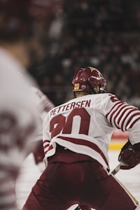 A hockey player in a red and white uniform is captured from behind, with the name 'Pettersen' and the number '20' displayed on the jersey. The player is in motion on the ice, holding a hockey stick, and wearing a helmet.