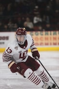 A hockey player in a white and maroon uniform is actively engaged on the ice, holding a hockey stick and poised for action. The player's gear includes a helmet, gloves, and skates, with a focused expression directed towards the game. The background shows a blurred audience and advertisements in an arena setting.