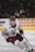 A hockey player in a white and maroon uniform is actively engaged on the ice, holding a hockey stick and poised for action. The player's gear includes a helmet, gloves, and skates, with a focused expression directed towards the game. The background shows a blurred audience and advertisements in an arena setting.