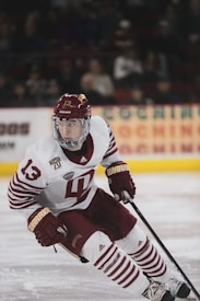 A hockey player in a white and maroon uniform is actively engaged on the ice, holding a hockey stick and poised for action. The player's gear includes a helmet, gloves, and skates, with a focused expression directed towards the game. The background shows a blurred audience and advertisements in an arena setting.