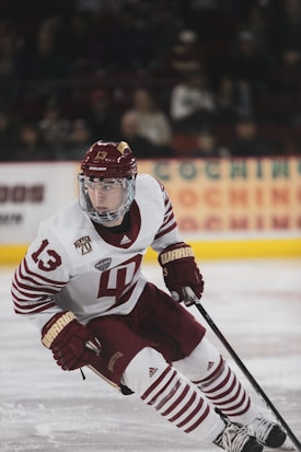 A hockey player in a white and maroon uniform is actively engaged on the ice, holding a hockey stick and poised for action. The player's gear includes a helmet, gloves, and skates, with a focused expression directed towards the game. The background shows a blurred audience and advertisements in an arena setting.