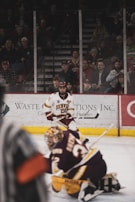 A hockey player from Denver in a white and maroon uniform is on the ice, positioned behind an opposing team’s goalie in black and maroon gear. The ice rink is surrounded by a crowd of spectators seated behind a clear protective barrier.
