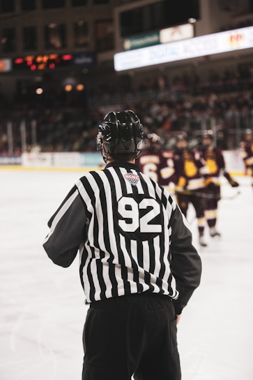 A hockey referee, wearing a black helmet and a striped jersey with the number 92, is standing on the ice rink. In the background, two teams of players in uniforms are visible, with figures blurred due to motion. Spectators fill the stands, which are also slightly blurred, indicating a lively scene.