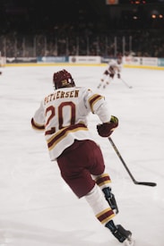 A hockey player in a maroon and white uniform skates across the ice, holding a stick. The jersey displays the name 'Pettersen' and the number 20. The arena is filled with spectators, and another player is visible in the background.
