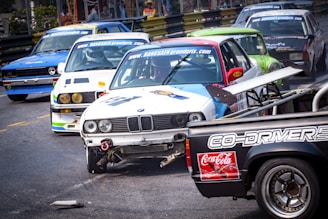 Several racing cars are on a track during a competitive event, with some vehicles showing visible damage. The scene is dynamic, with a close proximity between the cars suggesting intense competition.