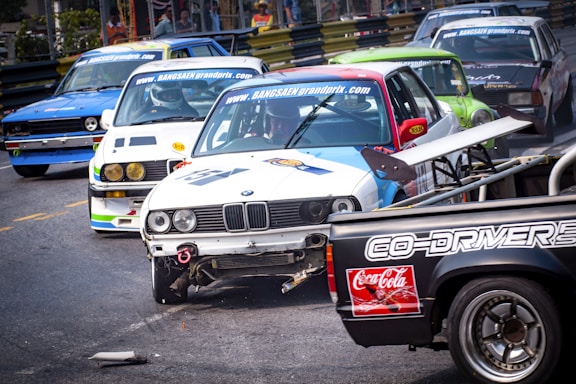 Several racing cars are on a track during a competitive event, with some vehicles showing visible damage. The scene is dynamic, with a close proximity between the cars suggesting intense competition.