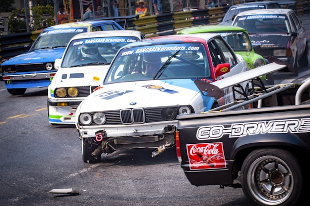 Several racing cars are on a track during a competitive event, with some vehicles showing visible damage. The scene is dynamic, with a close proximity between the cars suggesting intense competition.