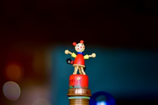 A colorful wooden toy sits on top of a cylindrical base. The toy resembles a small character with a round head, painted face, and extending arms. It features a red body and a blue collar, displaying a cheerful expression.