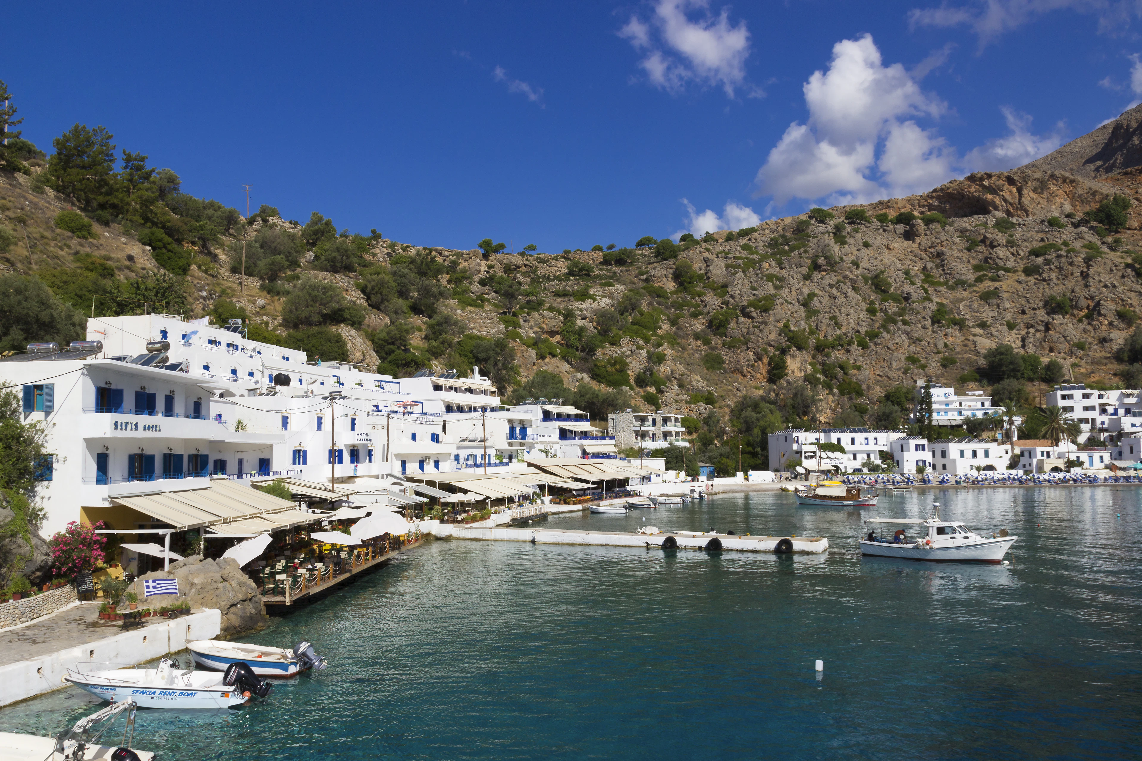 The whitewashed village of Loutro stacked around its horseshoe harbour on Crete's south coast, awnings over the waterfront tavernas and small boats moored on glassy turquoise water below bare brown mountains.