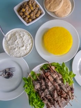 A meal consisting of skewered grilled meat served on a plate with fresh green herbs. Accompanying dishes include a plate of vibrant yellow saffron rice, a bowl of white sauce or dip with herbs, a bowl of seasoned olives, and several pieces of flatbread. Cutlery is neatly placed on an empty plate.