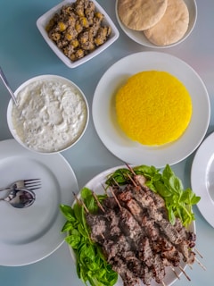 A meal consisting of skewered grilled meat served on a plate with fresh green herbs. Accompanying dishes include a plate of vibrant yellow saffron rice, a bowl of white sauce or dip with herbs, a bowl of seasoned olives, and several pieces of flatbread. Cutlery is neatly placed on an empty plate.