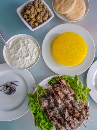 A meal consisting of skewered grilled meat served on a plate with fresh green herbs. Accompanying dishes include a plate of vibrant yellow saffron rice, a bowl of white sauce or dip with herbs, a bowl of seasoned olives, and several pieces of flatbread. Cutlery is neatly placed on an empty plate.